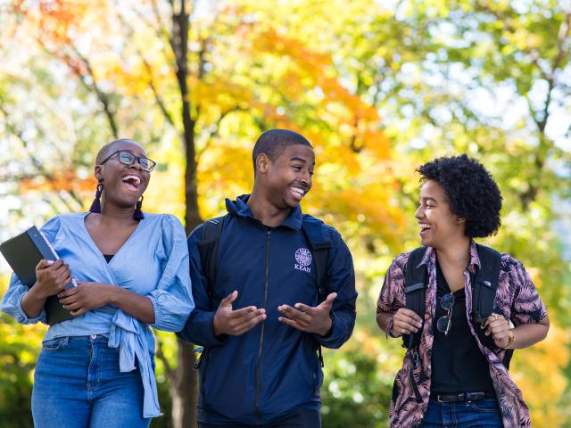 Three students laugh against a backdrop of fall foliage. 