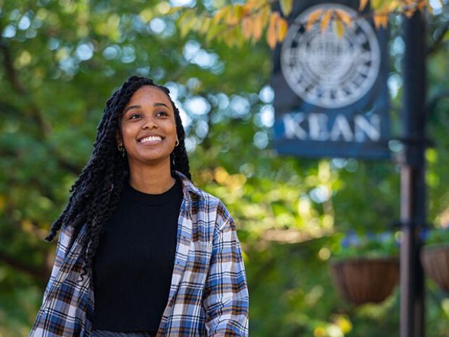 Student in plaid shirt walking outside with Kean flag in background