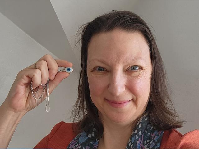 Smiling white female with brown hair, orange shirt and grey scarf holding up an evil eye amulet