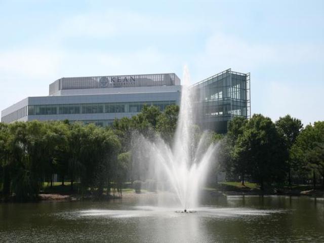 An active fountain in the middle of a small lake lined with trees; multi-level building with windows in the background.