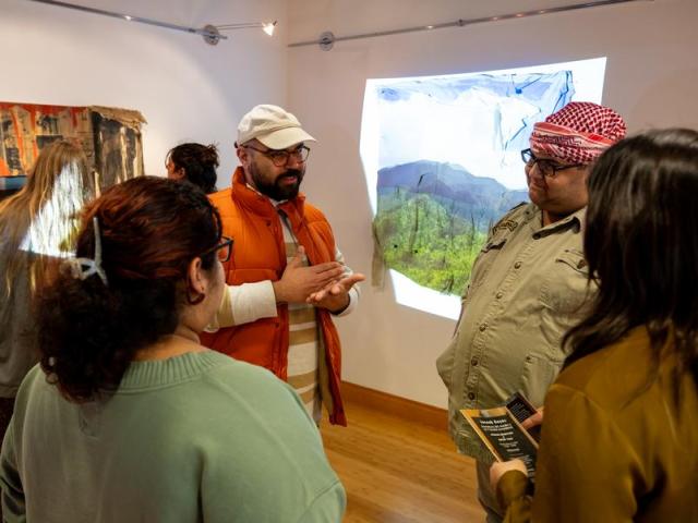 a man in a tan hat and orange vest talks to a group of three women in the middle of an art gallery