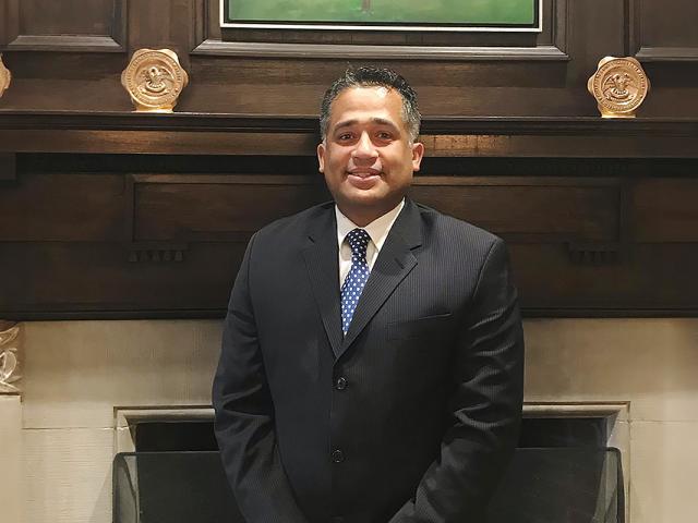 Alex Guzman, wearing a blue suit, stands in front of a fireplace.