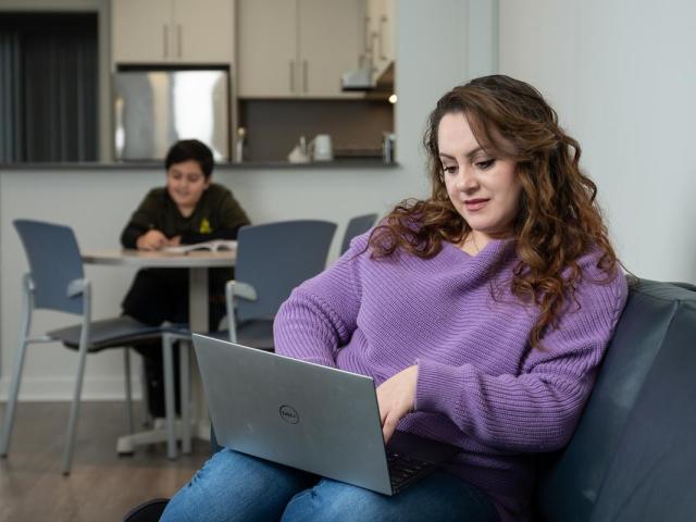 A woman works on her laptop. In the background, a young boy works at a table.