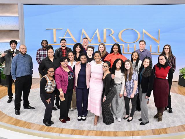 Group of students of diverse backgrounds standing with beside a woman in a pink dress, heels, short hair