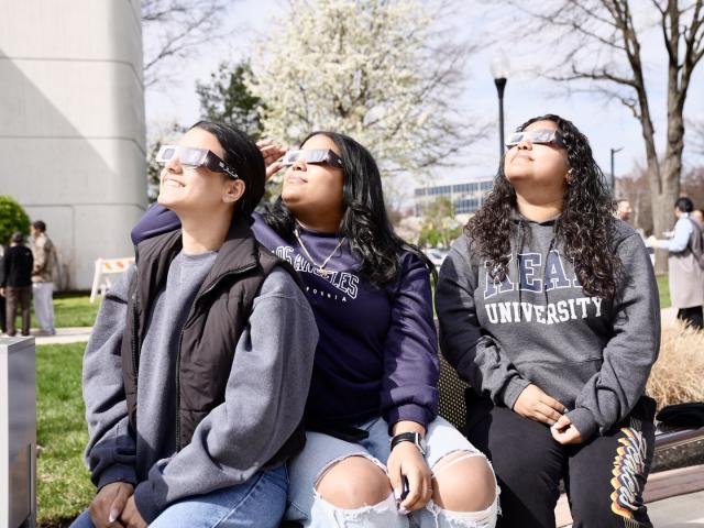 3 female of diverse background with solar eclipse glasses looking up with colorful clothing outside on campus
