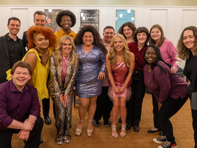 A diverse group of young men and women standing close to 3 adult women in sparkly outfits in the center