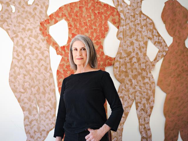 Woman with short gray hair wearing a black long-sleeved shirt standing in front of a mural painted with neutral-colored silhouettes of people
