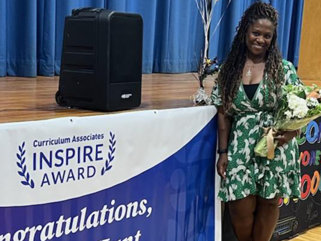 Tomeeko Hunt, a Black woman in a green floral dress, smiles as she stands next to an Inspire Awards Congratulations banner.