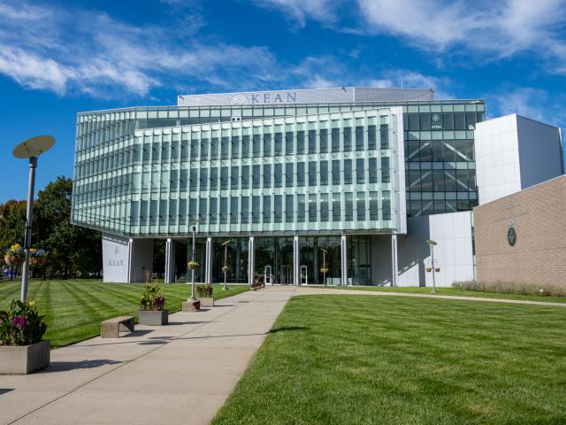 Outside of a large, six-story building lined with light green windows; green grass and blue skies.