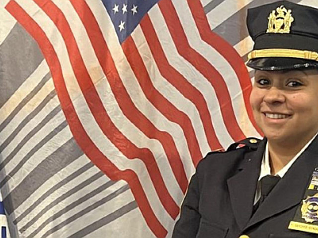 Smiling Hispanic woman in a black Port Authority uniform, including a hat, in front of a US flag