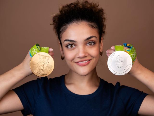 Smiling young Hispanic female holding two Olympic medals on either side of her face; she has short curly hair and is wearing a blue shirt.
