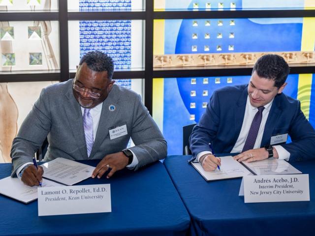 Black male wearing a gray suit and Hispanic male wearing a blue suit, side by side, each signing a formal document at a blue table; behind them is a stained glass window.