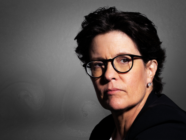 White female with short dark hair, black framed glasses, looking left, intently into the camara.