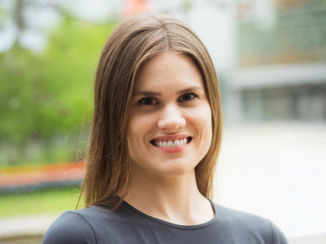Smiling white woman with light brown hair wearing a gray top; outdoor background is blurred.