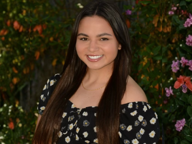 Smiling white female with long brown hair wearing a black top with yellow and white fllowers; blurred background of flowering trees.