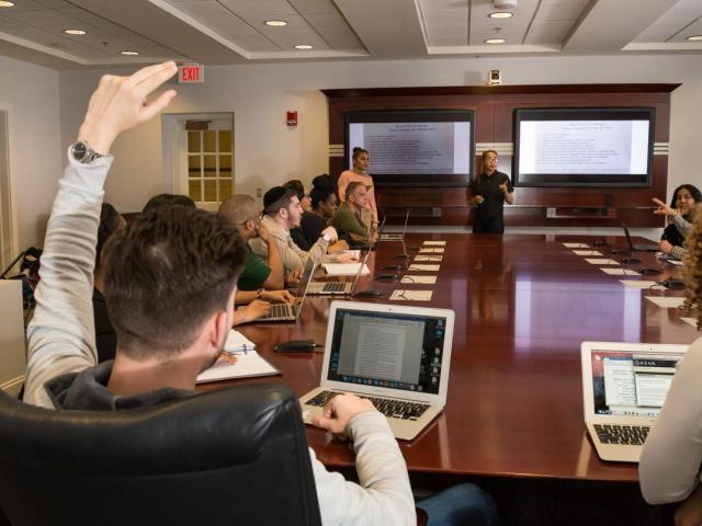 Students sitting around a large wooden conferenceroom table with laptops; one has their arm up.