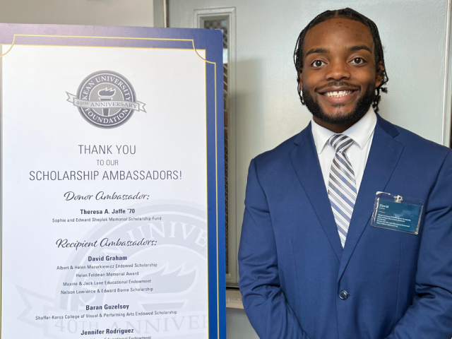 Smiling young Black male student in a blue suit and grey striped tie stands next to a sign announcing his scholarships.