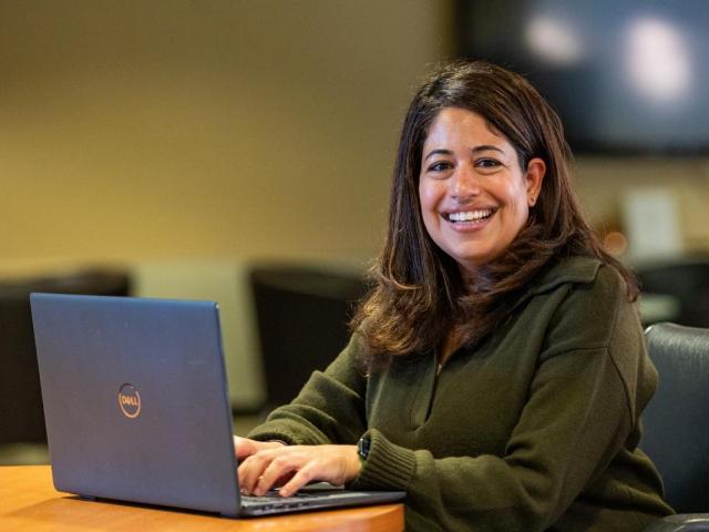 Smiling female with brown hair wearing a green sweater looks up from her laptop.