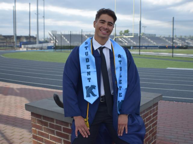 Man standing outside wearing graduation gown in front of athletic fields