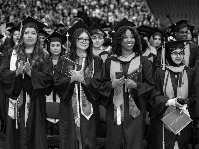 Four female students in blue caps and gowns, stand and applaud.