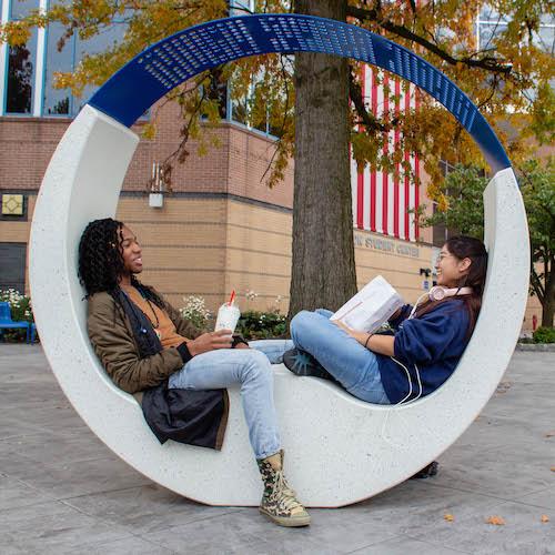 Two students seated on a sculptural bench outside on the Kean campus