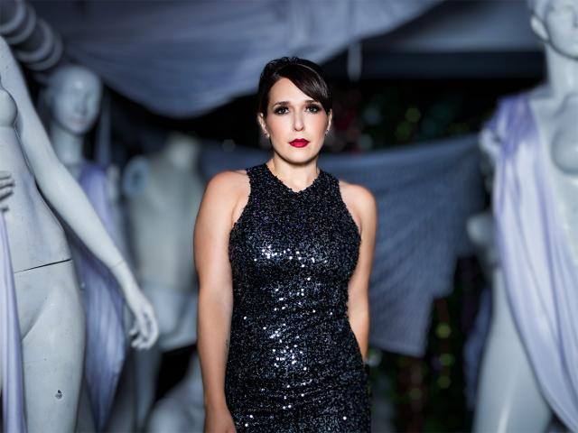Jessica Vosk standing with her hair tucked and sparkly tight dress and a background full of mannequins