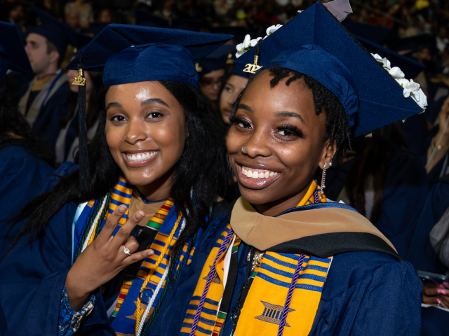 Two Black women, in Kean blue graduation caps and gowns, smile as they sit next to each other at Commencement.