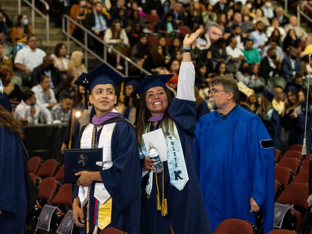 A woman in graduation cap and gown waves to someone in the audience.