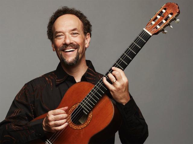 A white male with curly hair, and a mustache a big smile on his face and a classical guitar between his hands
