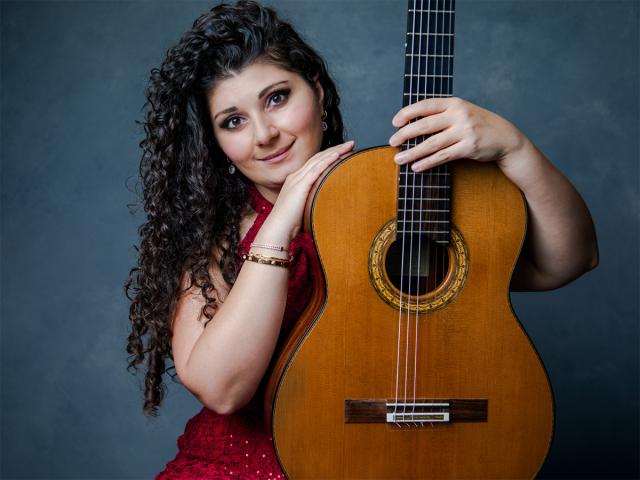 A woman with curly hair, wearing a red dress, affectionately holds a classical guitar against a soft gray backdrop. Her expression is serene, and her pose suggests a deep connection to the instrument. The guitar's warm wooden tones contrast with her elegant attire, emphasizing themes of music and artistry.