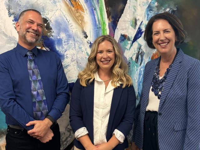 Three people standing in front of an expressive painting, smiling for the camera