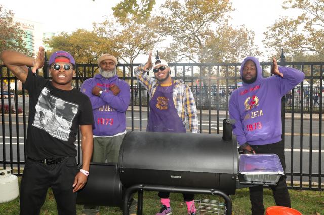 Four fraternity brothers pose for a picture in front of a grill holding up their fraternity hand signs. 