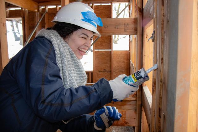 Female student holds caulking gun and caulks a house being built 