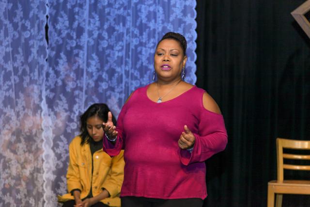 Woman on stage in bright, purple shirt acting out monologues of a victim of domestic violence.