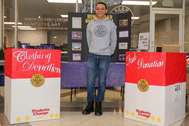 Roberto Adams poses in front of poster about homelessness with clothing and food drive boxes next to him