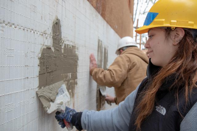 A student uses tools to stucco the side of a home.