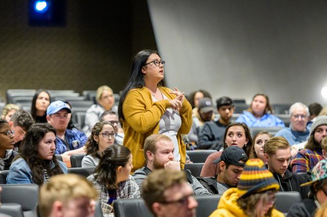 A female student stands up in the crowd and asks a question to the speaker with microphone in hand 