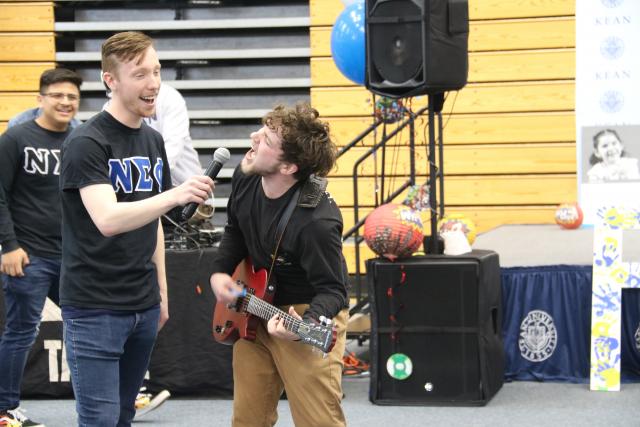 Two students perform on stage as one plays guitar and the other holds the microphone up for him excitedly. 