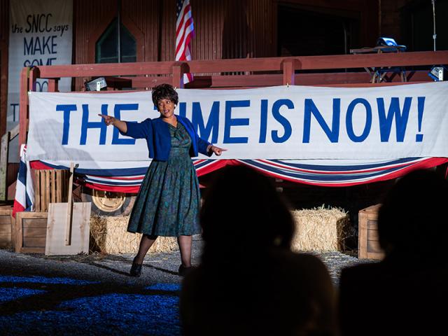 An African-American woman stands in front of banner that reads The Time is Now