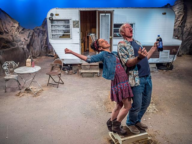 A young woman and an older man (sing?) in the desert in front of an RV