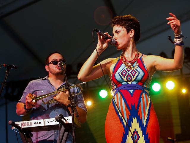  Hispanic Female with short brown hair and a multicolored dress singing on stage with a man playing the trumpet.