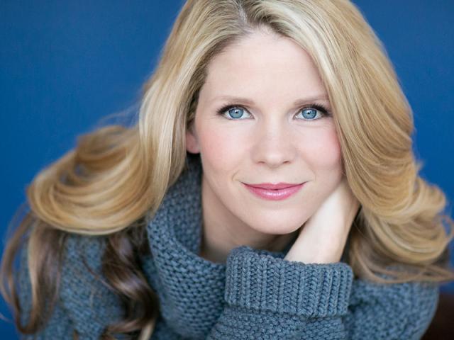 Blonde hair, blue eyed white woman wearing a blue knit sweater.