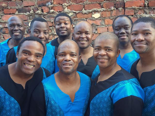 A group of Black men standing infront of a brick wall wearing balck and blue African costumes.