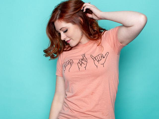 Red-haired white woman in blue jeans and pink shirt with the ASL letters that spells out “Try”