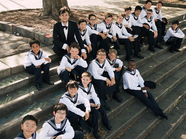 The choir is sitting on stairs with their mentor, under a tree