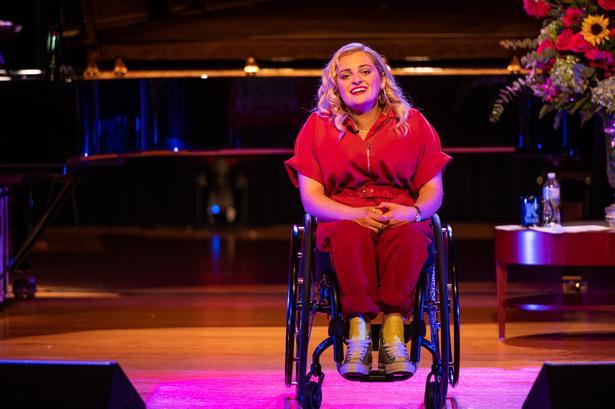 Ali Stroker, a female with curly blond hair, sitting in her wheelchair on stage. She is wearing a red jump suit. Head tilted, singing. Stage is lit pink. Piano and flowers can partially be seen behind her.