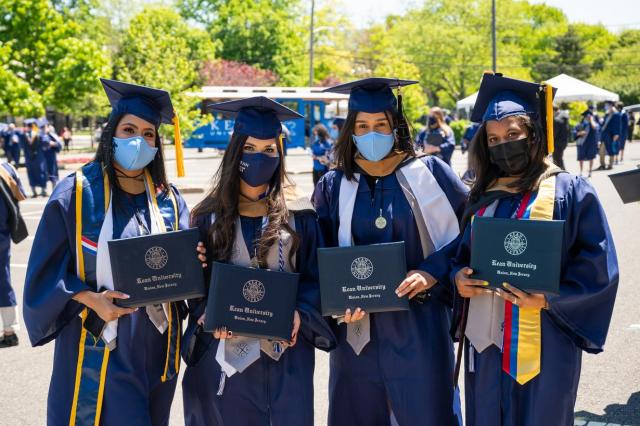 Four Kean graduates wearing masks and commencement regalia pose with their diplomas.