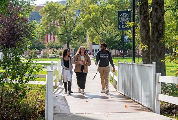 Three students, two women and a man, walk over a white bridge on Kean University's Union campus