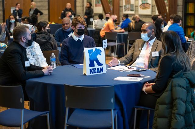 Five people of different ethnicities and races seated at a table with a Kean logo as the center piece, dressed in suits and talking to each other.