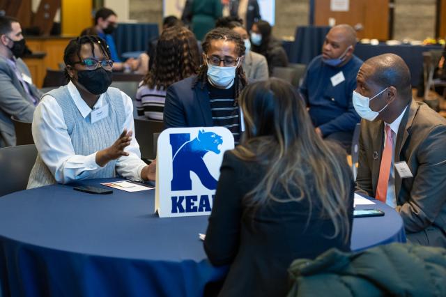 Four people of different ethnicities and races seated at a table with a Kean logo as the center piece, dressed in suits and talking to each other.